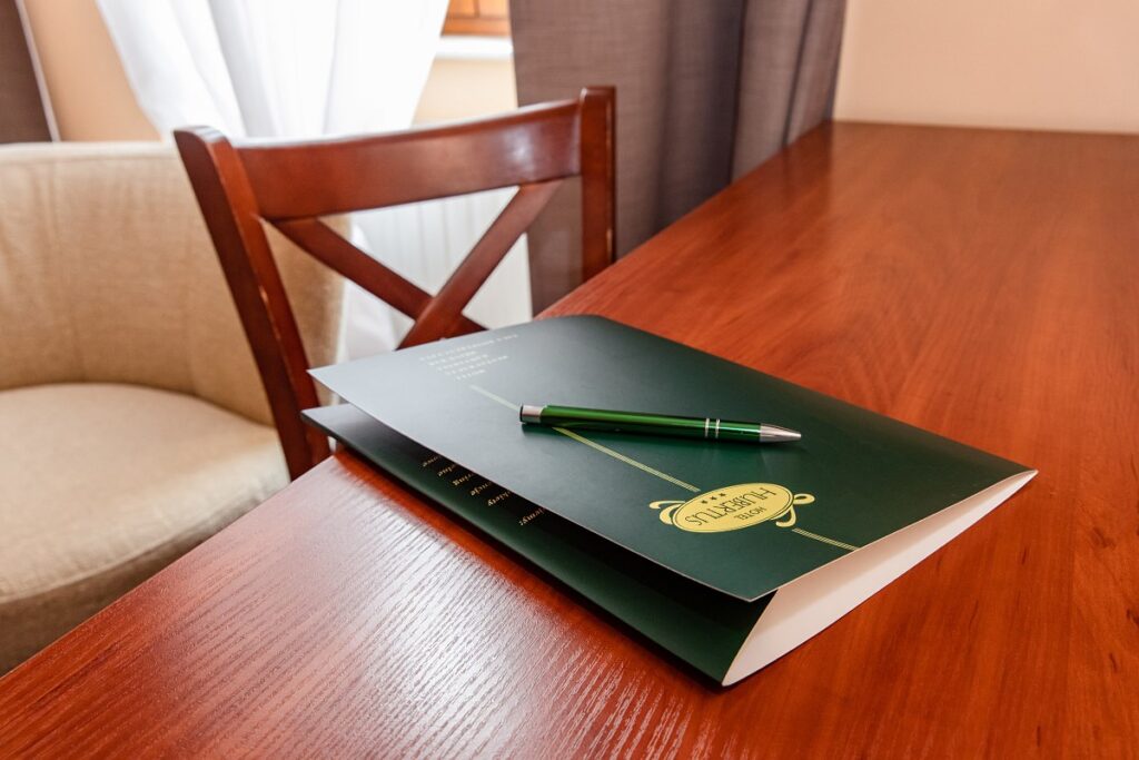 Notatnik z zieloną okładką i długopisem na drewnianym stole obok krzesła, przy jasnym oknie.