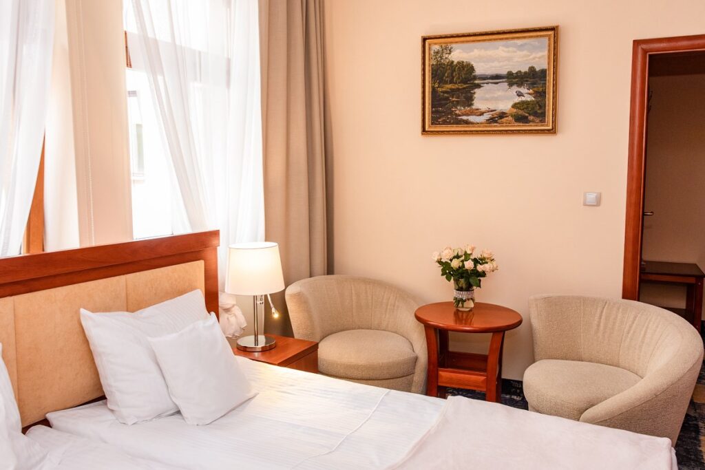 Przytulny pokój hotelowy z łóżkiem, lampką nocną, fotelami i bukietem kwiatów na stoliku.