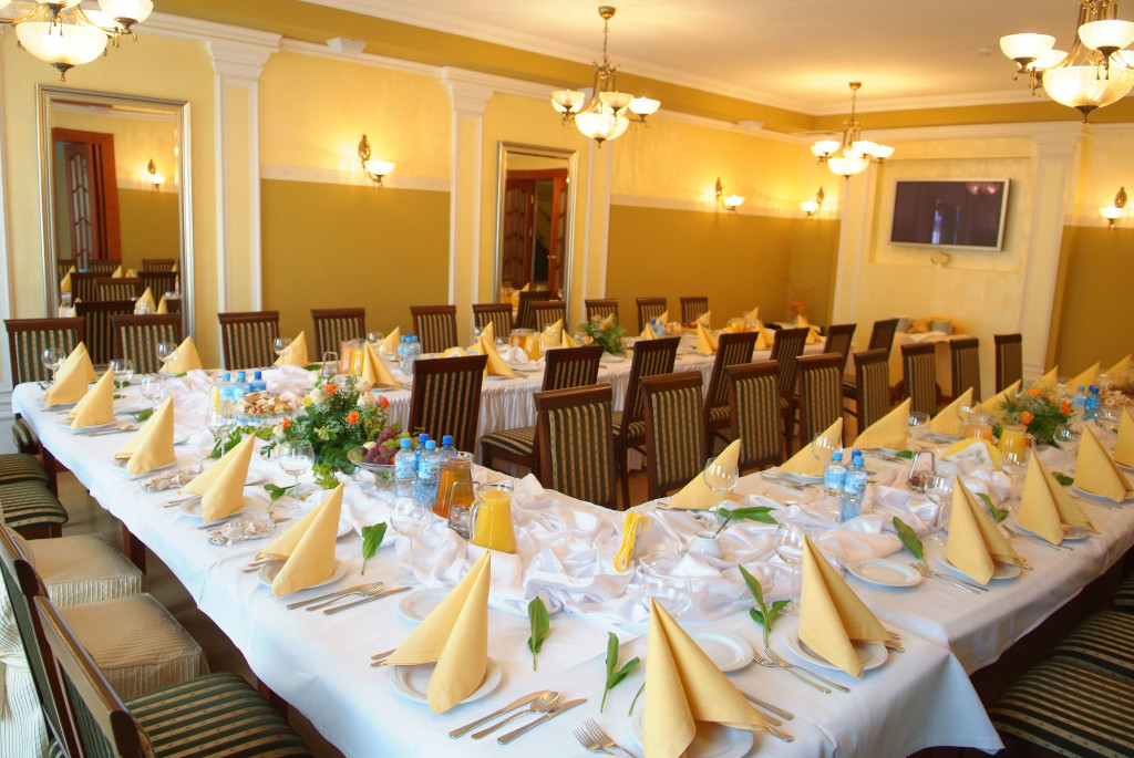 Elegancka sala bankietowa z zastawionymi stołami, serwetki w złotym kolorze, kwiatowe dekoracje i nastrojowe oświetlenie.
